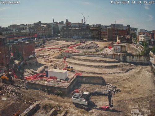 Construction site in an urban area with terraced excavation levels, machinery, and red safety barriers. Surrounding the site are older brick buildings and cranes in the background under a clear blue sky.