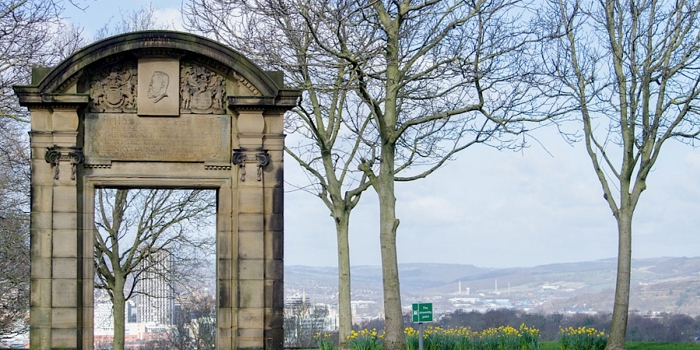 A historic stone archway with decorative carvings stands in a park surrounded by bare trees. The arch frames a distant view of a cityscape with hills in the background. Yellow daffodils bloom in the grassy area near the arch, and a small green sign is visible on the lawn. The sky is bright with scattered clouds, creating a clear and open atmosphere.