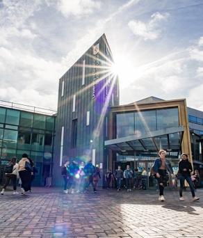 A modern building with a glass facade and a tall, narrow central structure. The sun shines brightly, creating lens flares and shadows. Several people are walking or standing near the entrance. The sky is partly cloudy, contributing to the bright atmosphere.