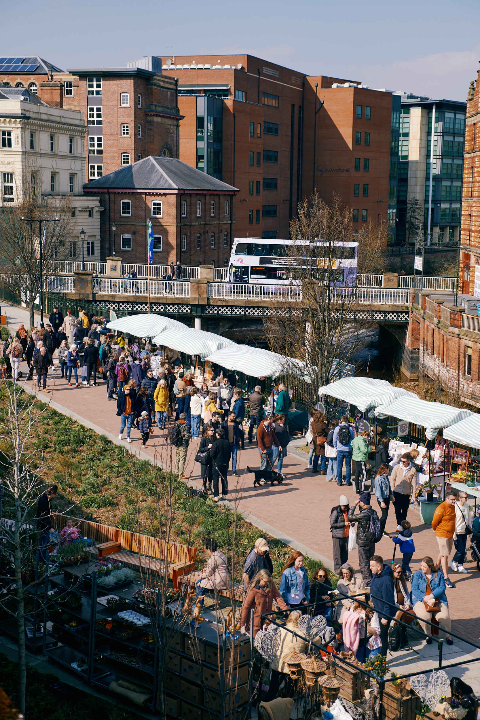 An open air market in a city centre, next to a river. There are lots of people walking about and browsing the stalls.