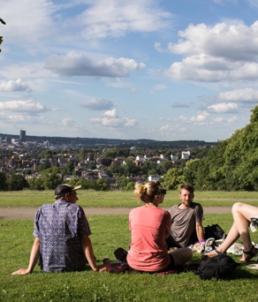 A group of four people, sat in Meersbrook Park, enjoying a sunny day. One is sat in a folding chair and the other three are sat on the grass.