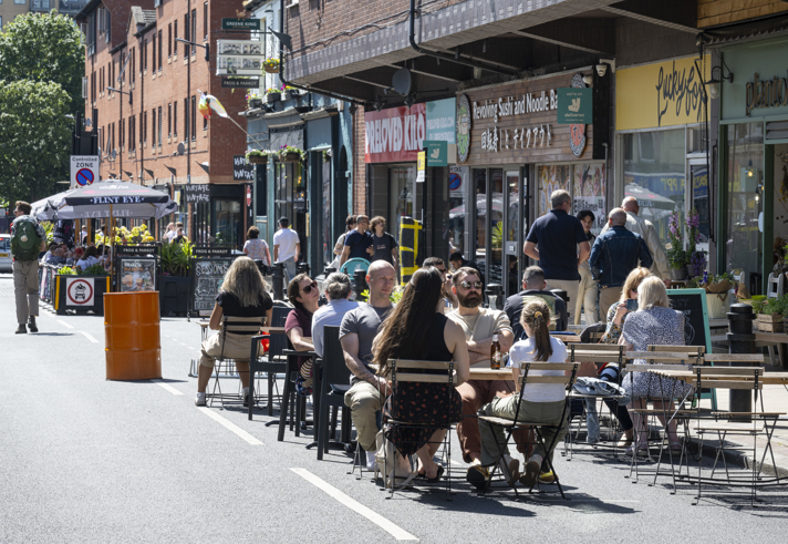 People are seated at outdoor tables on a closed-off city street, enjoying food and drinks in sunny weather. The street is lined with shops and restaurants, including signs for a noodle bar and a café. A bright orange barrel and potted plants decorate the area, while pedestrians walk along the street in the background. The scene conveys a lively, social atmosphere in an urban setting.