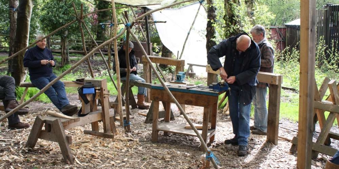 People working at wooden benches in an outdoor woodland workshop, using hand tools under a simple canopy surrounded by trees.