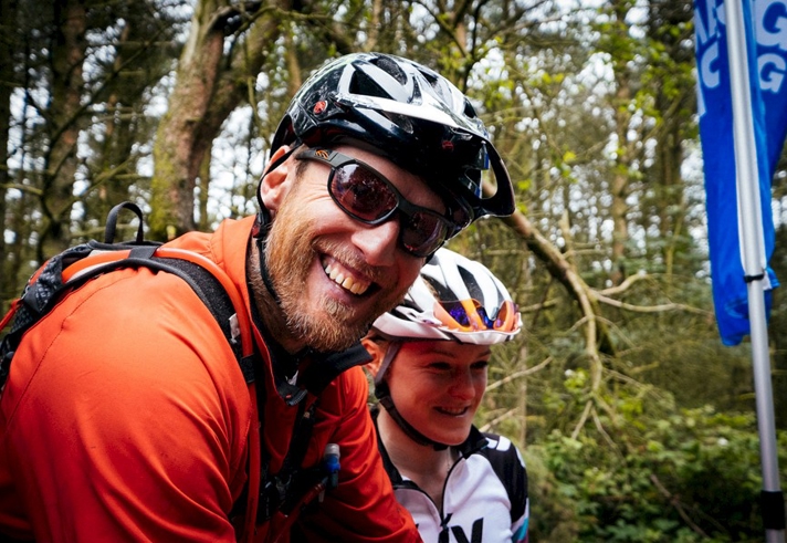 Male cyclist in sunglasses and cycling helmet smiling at the camera with his child.