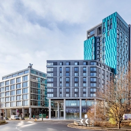 Modern cityscape featuring two contemporary high-rise buildings at a roundabout. The left building has a glass façade with a grid-like design, while the taller building on the right has a striking blue and gray exterior with geometric patterns. Bare trees frame the scene, and signage at street level reads “New Era Square.” The sky is partly cloudy, creating a bright and open atmosphere.