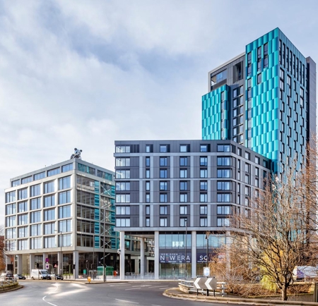 Modern cityscape featuring two contemporary high-rise buildings at a roundabout. The left building has a glass façade with a grid-like design, while the taller building on the right has a striking blue and gray exterior with geometric patterns. Bare trees frame the scene, and signage at street level reads “New Era Square.” The sky is partly cloudy, creating a bright and open atmosphere.
