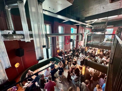 Interior view of a busy bar or restaurant with an industrial-style design, featuring exposed metal pipes, high ceilings, and large windows letting in natural light. The space is filled with people standing and sitting at tables, socializing and enjoying drinks. Warm lighting from wall lamps and hanging fixtures adds to the lively atmosphere.
