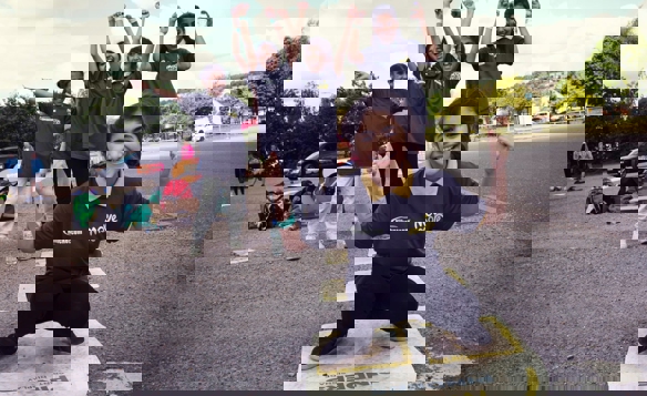 A group of children wearing matching dark gray “Move More” t-shirts participate in an outdoor activity on a paved area. One child is crouching in the foreground on a circular marker with text, while others stand behind with arms raised in celebratory poses. Sports bags and equipment are visible on the ground, and trees and parked cars can be seen in the background under a partly cloudy sky.