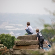 Two children are climbing and sitting on large stacked stone slabs in a natural outdoor setting. The background shows a hazy view of a distant town or city with trees and greenery in the foreground.