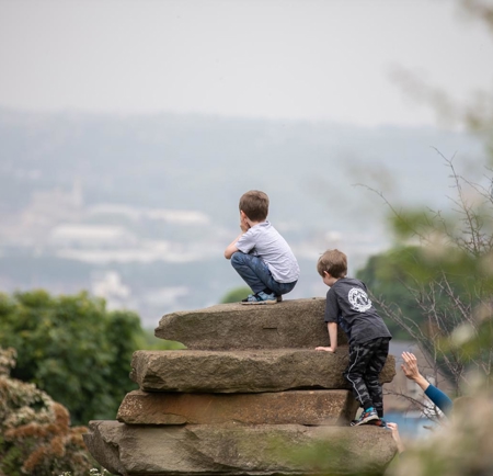 Two children are climbing and sitting on large stacked stone slabs in a natural outdoor setting. The background shows a hazy view of a distant town or city with trees and greenery in the foreground.