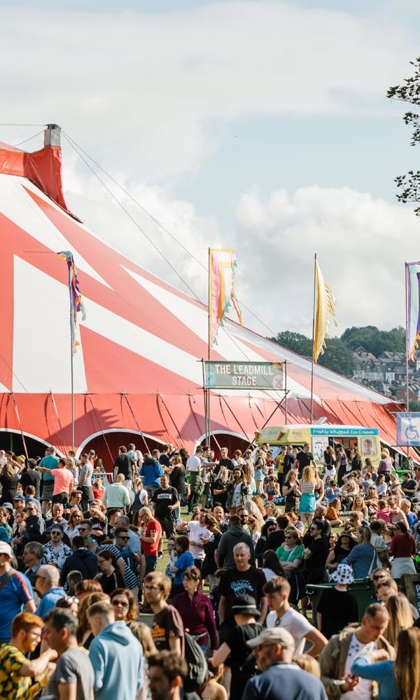 Large outdoor festival crowd gathered near a big red-and-white striped tent, with colourful flags and trees in the background under a partly cloudy sky.