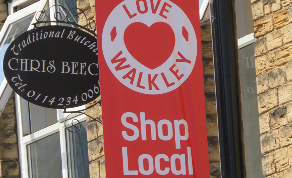 Close-up of a red banner attached to a lamppost with the words “LOVE WALKLEY” and “Shop Local” in white text, featuring a heart symbol. Next to it is a black oval sign for a traditional butcher shop reading “Chris Beec…” with a phone number partially visible. The background shows a stone building with a window.