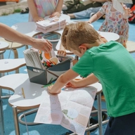 Child leaning over a table with a coloring sheet, surrounded by other children and adults at an outdoor craft activity. The table holds a box of colored pencils and markers, and several stools are arranged on a bright blue surface.