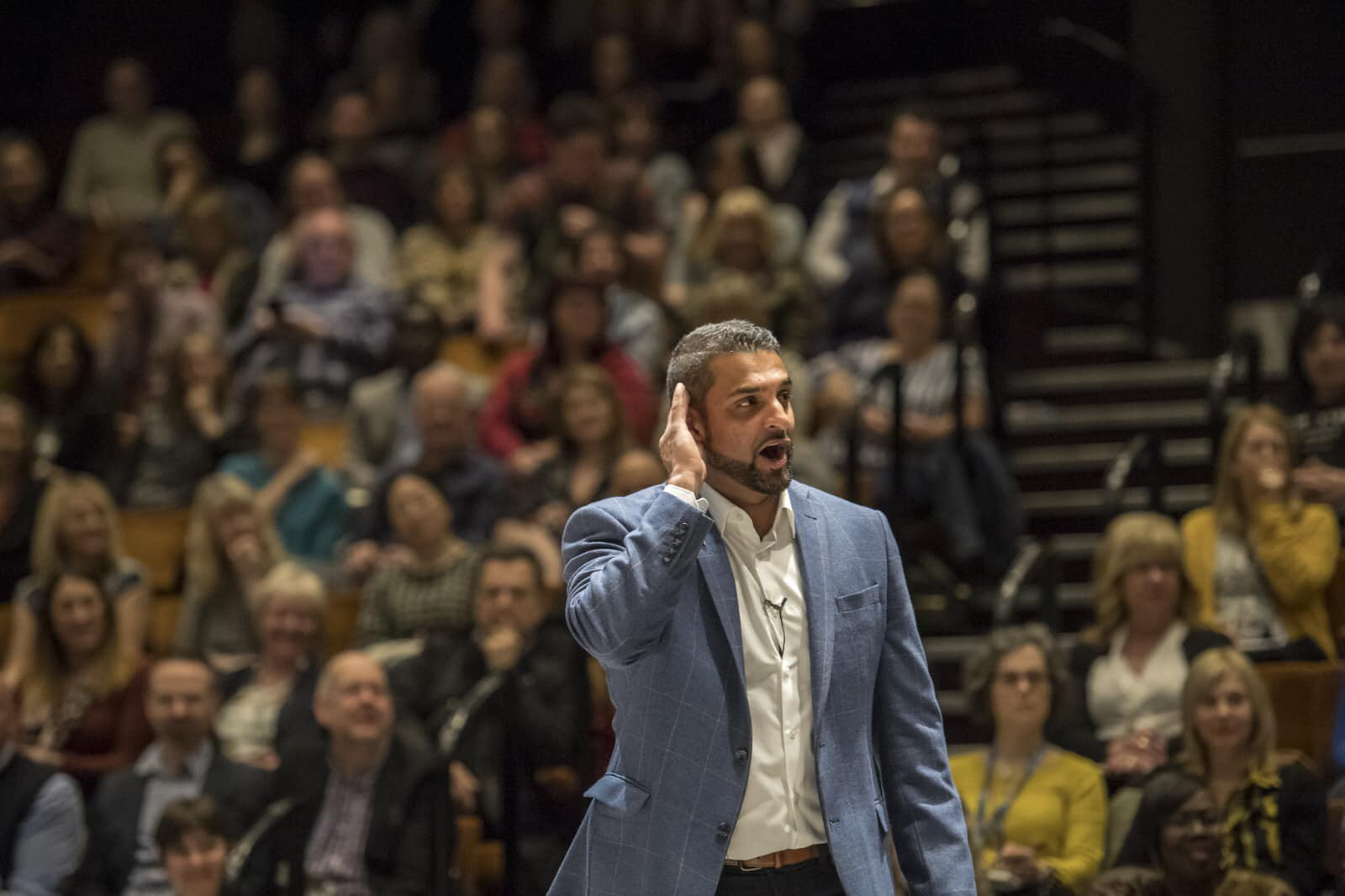 Person in a blue blazer standing in front of a large seated audience in an auditorium. The individual is holding one hand near the ear, possibly listening or engaging with the crowd. The background shows rows of people seated on tiered benches, with steps and railings visible on the right side. Lighting is focused on the person in the foreground, creating a contrast with the dimmer audience area.