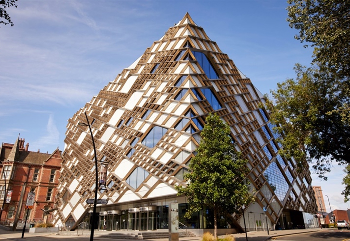 Modern pyramid-shaped building with a striking geometric design featuring glass panels and wooden lattice framework, located on a street corner with trees and older brick buildings nearby under a clear blue sky.