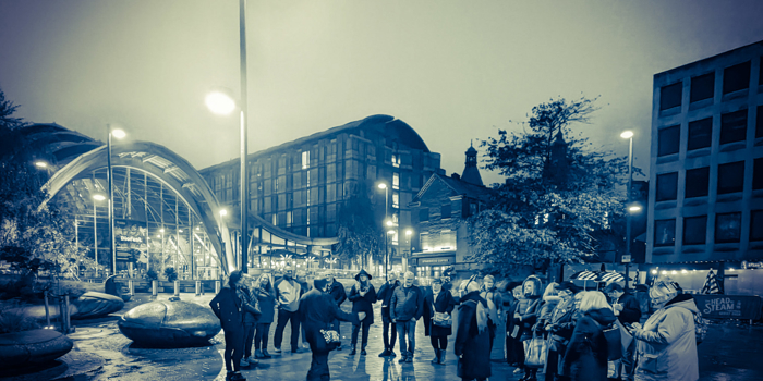 A group of people gathered round a tour guide, in Tudor Square in the centre of Sheffield, at night.