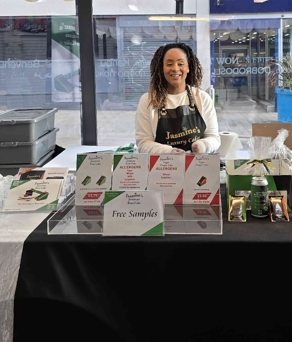 A display table set up for a food sampling event inside a bright indoor space with large glass windows. The table is covered with a black and white cloth and features neatly arranged packaged snacks, promotional signs, and a tray labeled “Free Samples.” There are clear acrylic stands holding additional products on the right side, and a small green sign with food hygiene rating on the left.