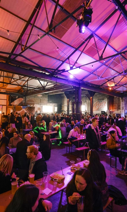 Large indoor venue with high industrial-style ceiling and exposed beams, filled with people seated at long wooden tables and standing in groups. Warm orange and purple lighting creates a lively atmosphere, with food and drinks visible on tables. A bar and food stalls are in the background.