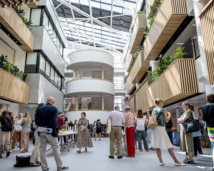 The Heartspace in Hallam University: Bright, modern atrium with a glass roof and natural light. People are gathered for an event, standing near tables with refreshments. The space features multiple levels with wooden slatted balconies, green plants, and white curved staircases in the centre, creating an open and airy atmosphere.