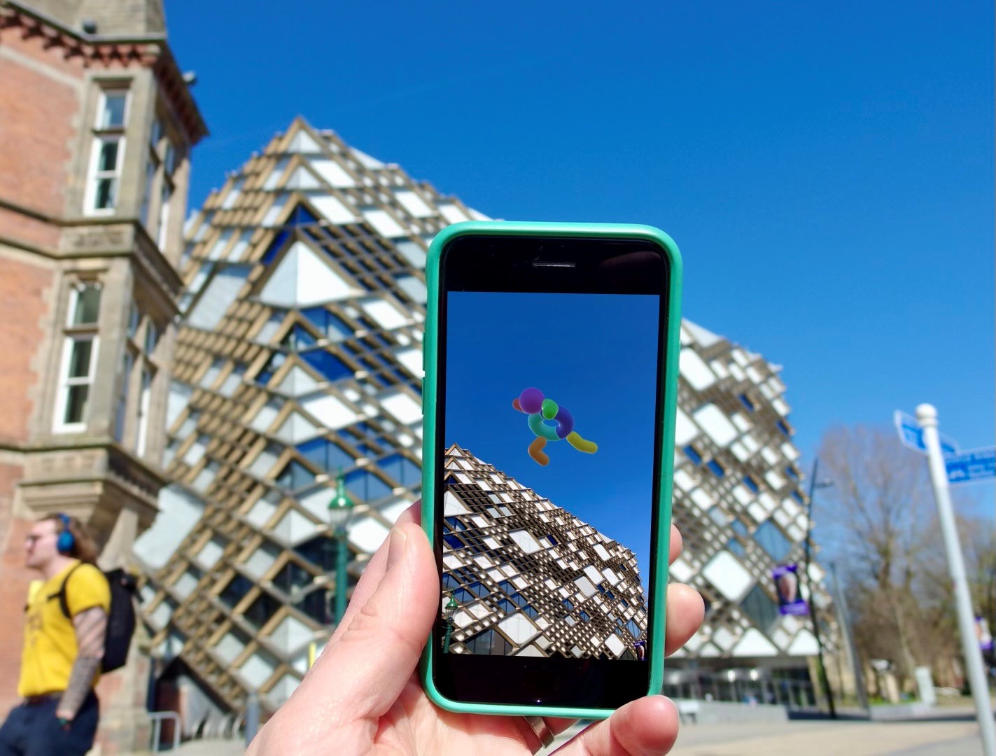 Hand with mobile phone held up in front of The Diamond Building, Sheffield, showing  Look Up AR art 