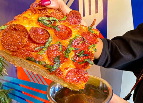 Close-up of a large slice of pepperoni pizza topped with fresh basil leaves, held above a metal dipping cup filled with sauce. The background features a colorful wall with bold geometric patterns in blue, red, and white.