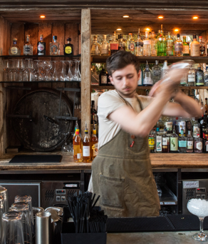Bartender behind a rustic wooden bar shaking a cocktail shaker. The bar is stocked with various bottles of spirits, wine, and mixers arranged on shelves, along with glassware and a large wooden barrel in the center. The countertop has cocktail-making tools, straws, and an empty glass with ice ready for a drink. Warm lighting creates a cozy atmosphere.
