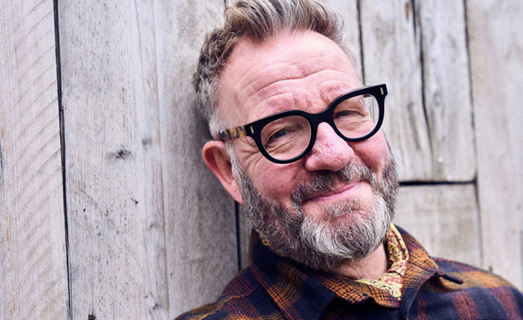 The artist Pete Mckee leaning against a wooden fence, smiling at the camera.